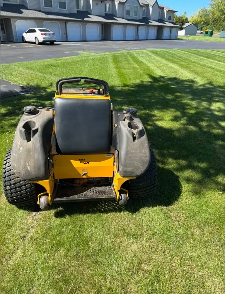 Overhead view of a yellow and black zero-turn radius mower on a striped lawn with suburban houses and garage in background