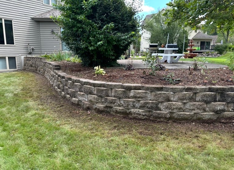 Residential backyard with curved stone retaining wall, mulched garden bed, shrubs, and green lawn in front of a house