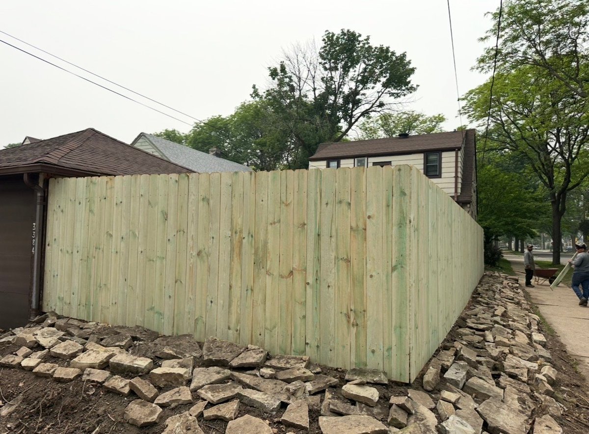 Weathered wooden fence with peeling green paint along a residential street, with buildings and trees visible in background